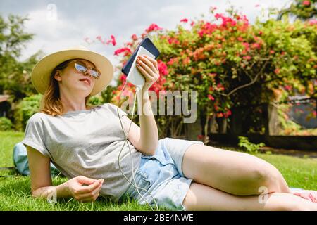 Une femme s'est couponée sur l'herbe verte, tenant le téléphone mobile et la banque d'alimentation portable au pique-nique dans le parc Banque D'Images