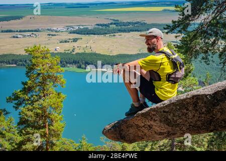 Coureur d'athlète reposant sur le bord d'une falaise qui s'y colle. Athlète barbu en short et T-shirt jaune. Vue sur le lac. Sports de plein air. Lac d'Imantau Banque D'Images