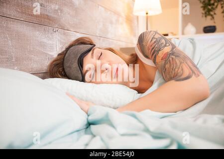 Belle femme avec tatouage dormant dans le lit à la maison avec masque de couverture. Banque D'Images