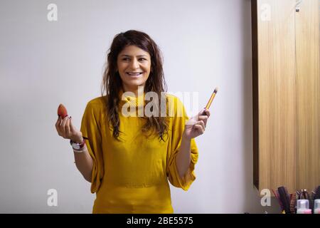 Jeune femme souriant avec un mixeur de maquillage dans une main et une brosse dans une autre. Banque D'Images