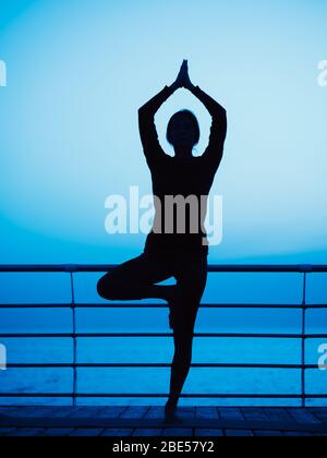 Jeune fille mince pratiquant le yoga sur le remblai contre l'océan ou la mer à l'heure de nuit. Silhouette de femme dans asana de l'arbre. Concept de somnambulisme Banque D'Images