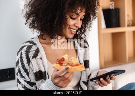 Image de la jeune femme africaine souriante à l'intérieur à la maison manger de la pizza à l'aide du téléphone portable. Banque D'Images