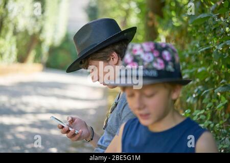 Deux frères adolescents en vacances d'été, dont l'un tient un téléphone mobile assis sous les arbres dans la verdure d'un parc Banque D'Images