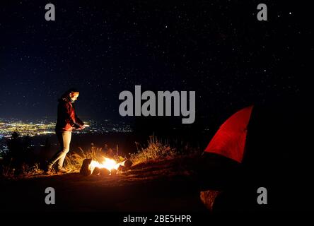 Belle fille randonneur ses mains chaudes près de camp sous ciel de nuit avec les étoiles et les lumières de la ville à l'arrière-plan. Banque D'Images