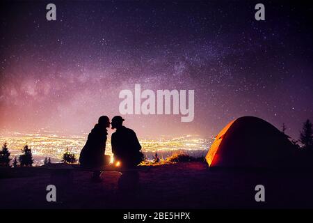 Couple heureux en silhouette embrassant près de camp et tente d'orange. Ciel nocturne avec voie lactée les étoiles et les lumières de la ville, à l'arrière-plan. Banque D'Images