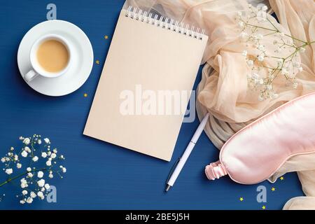 Bureau féminin avec bloc-notes en papier vierge, tasse à café, masque rose pour les yeux de sommeil, fleurs. Maison confortable, concept Happy Morning. Plat, vue du dessus Banque D'Images