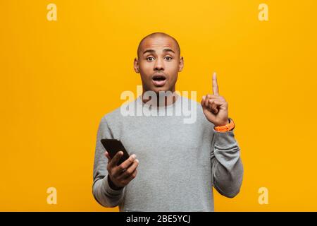 Portrait d'un jeune homme africain enthousiaste et décontracté, se tenant sur fond jaune, tenant le téléphone mobile, idée Banque D'Images