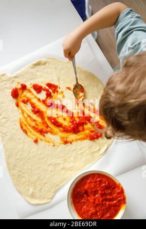 Petit garçon mettant la tomate avec une cuillère sur une pâte à pizza Banque D'Images