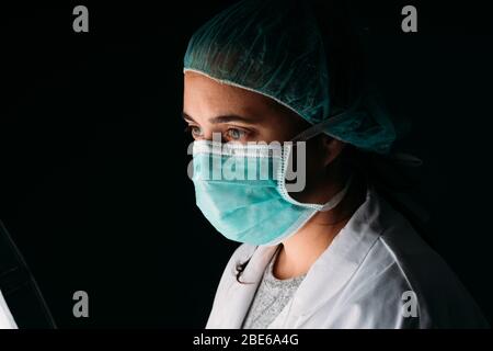 Jeune femme latérale médecin portant un masque chirurgical médical, une casquette et des vêtements de protection contre les virus sur fond noir avec espace de copie. COVID-19 le coronavirus Banque D'Images