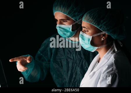 Portrait étroit de deux médecins de deux côtés de femme et d'homme travaillant avec le masque chirurgical médical, la casquette médicale et les vêtements de protection contre les virus sur fond noir Banque D'Images