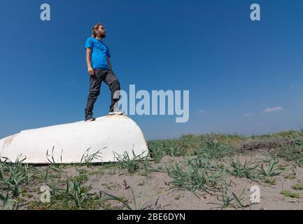 Un homme est debout sur un bateau blanc sur le sable. Il y a des plantes dans le sable. Le ciel est clair et bleu. Banque D'Images