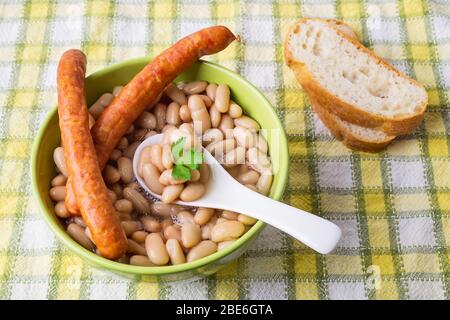 Haricots blancs, deux saucisses fines et une cuillère en céramique dans un bol vert près de tranches de pain sur une nappe à damier. Source de protéines végétales. Banque D'Images