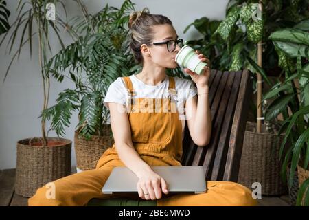 Femme jardinier européen portant des combinaisons, se reposant après le travail, assis sur une chaise en bois dans la serre à la maison, tenir réutilisable café/thé mug, à côté Banque D'Images