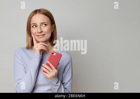 Studio portrait d'agréable gaie belle femme européenne regarde l'espace vierge de copie, tenant smartphone, isolé sur fond beige. Positif Banque D'Images