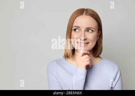 Joyeuse femme européenne avec des cheveux courts, vêtu d'un chemisier de lavande, touche le menton et se met à côté d'un espace de copie vide pour le produit, en pensant au shoppi Banque D'Images