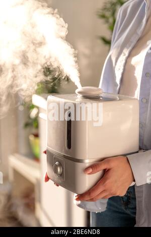 Femme tenant dans ses mains l'humidificateur d'huile d'arôme de travail dans la pièce ensoleillée. Humidification de l'air dans l'appartement pendant la période de l'auto-isolation Banque D'Images