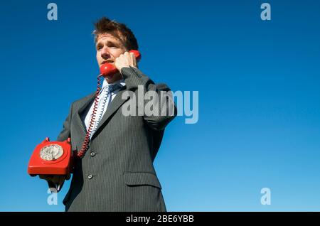 Homme d'affaires ayant une conversation urgente sur un téléphone rotatif rouge à l'ancienne debout à l'extérieur dans le ciel bleu ensoleillé Banque D'Images