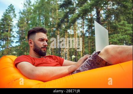 L'homme barbu travaille et souriait avec un ordinateur portable dans le parc sous l'arbre. Un travailleur indépendant heureux est assis et utilise l'application ou le site Web sur l'herbe. Travail Banque D'Images