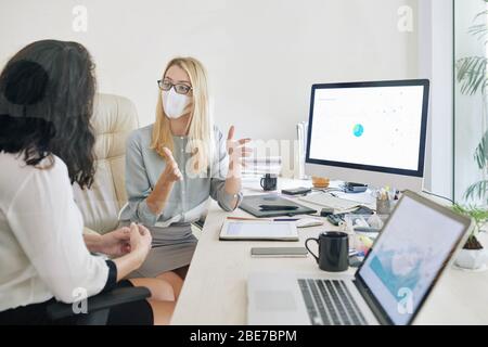 Jeune femme d'affaires dans des lunettes et masque médical gestuelle activement lors de l'explication de l'idée à la colesgue Banque D'Images