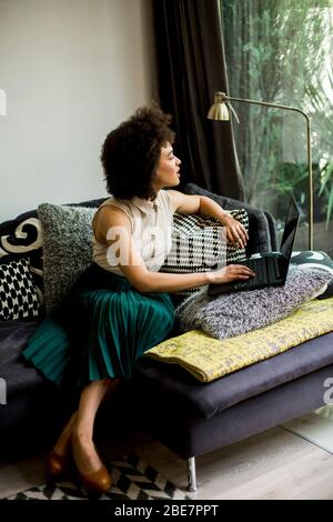 Jolie jeune fille avec des cheveux bouclés sur l'ordinateur portable tout en s'asseoir sur le canapé à la maison Banque D'Images