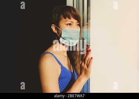 Une femme asiatique ennuie portant un masque de visage regardant la fenêtre pendant la quarantaine à la maison Banque D'Images