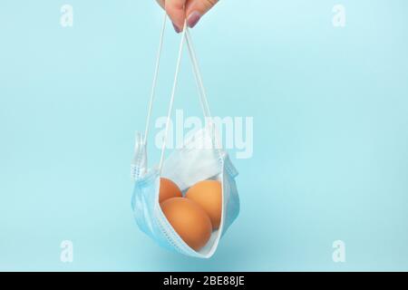 La main des femmes en gros plan contient un masque de protection chirurgical avec des œufs de Pâques sur un fond bleu. Vacances pendant la pandémie de coronavirus et la quarantaine. Banque D'Images