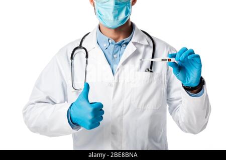 vue rognée du médecin dans le masque médical et le manteau blanc tenant le thermomètre numérique et montrant le pouce isolé sur blanc Banque D'Images