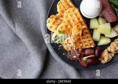 Un copieux petit déjeuner sur une plaque sombre sur le lit avec couvre-lit gris dans la chambre de l'hôtel. Vue de dessus. Service d'étage. Gaufres croustillantes avec confiture, œufs durs, poivre Banque D'Images