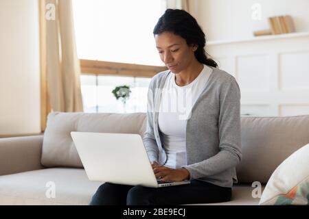 Une jeune fille noire sereine s'assoit sur un canapé et travaille sur un ordinateur portable Banque D'Images