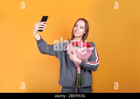 Bonne jeune femme avec des fleurs roses prenant selfie sur jaune arrière-plan Banque D'Images