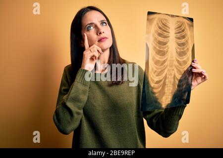 Jeune femme brunette avec des yeux bleus tenant des rayons X des poumons sur fond jaune sérieux visage pensant à la question, idée très confuse Banque D'Images