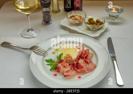 Fromage de chèvre chaud au miel et au bacon servi sur un plat blanc. Rosettes aux amandes et aux olives vertes et un verre de vin blanc sur la table. Banque D'Images