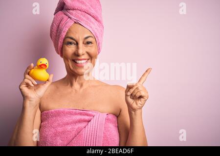 Femme d'âge moyen portant une serviette de bain rose de soins du corps de beauté tenant le canard jaune en caoutchouc très heureux pointant avec la main et le doigt sur le côté Banque D'Images