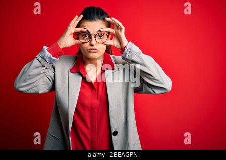 Jeune femme d'affaires brunette belle portant une veste et des lunettes sur fond rouge essayant d'ouvrir les yeux avec les doigts, endormie et fatigué pour le matin f Banque D'Images