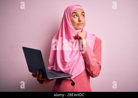 Jeune femme d'affaires belle portant le hijab musulman rose et la veste d'affaires utilisant un ordinateur portable sérieux face à la question, très confus idée Banque D'Images