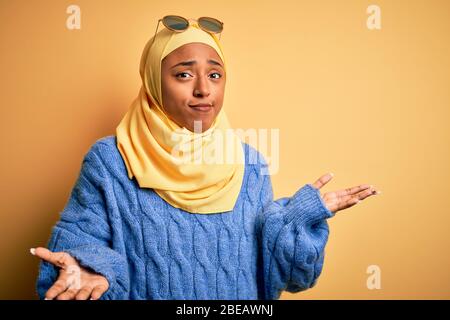 Une jeune étudiante africaine américaine portant un hijab musulman jaune et des lunettes de soleil se fait sans indice et confus avec les bras et les mains relevés. Doute con Banque D'Images