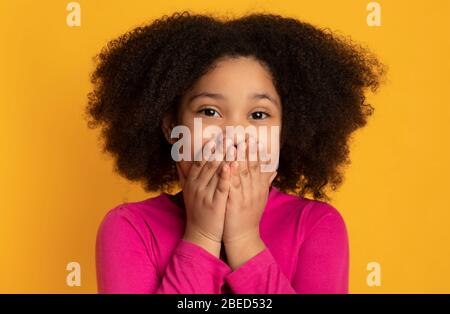 Jolie petite fille noire couvrant la bouche avec les mains sur fond jaune Banque D'Images