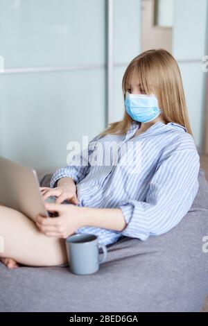 Jeune fille dans un masque médical, une pyjama et une tasse de café assis sur un beanbag à la maison en isolement. Concept d'étude à distance ou travail indépendant du Banque D'Images