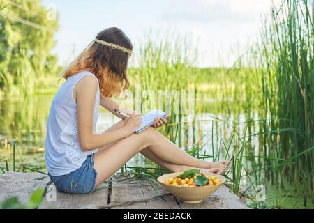 Belle fille romantique adolescent assis sur le pont près de l'eau, fille écrit dans des expériences personnelles d'ordinateur portable, bol de cerises, profitant de la beauté de pict Banque D'Images