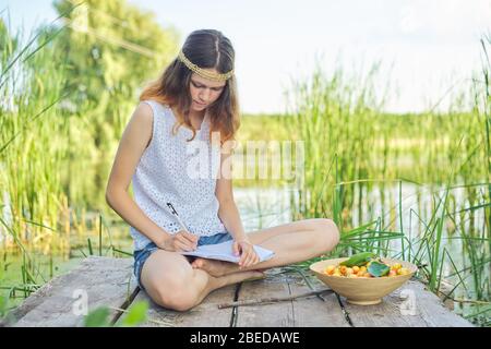 Belle fille romantique adolescent assis sur le pont près de l'eau, fille écrit dans des expériences personnelles d'ordinateur portable, bol de cerises, profitant de la beauté de pict Banque D'Images