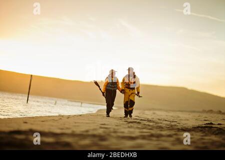 Happy senior couple marche main dans la main tout en portant des pagaies le long d'une plage de sable. Banque D'Images