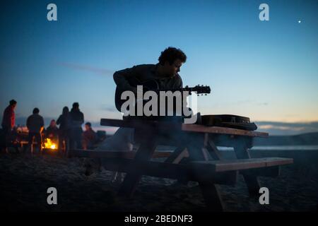 Adolescent jouant de la guitare sur une table de pique-nique à la plage au coucher du soleil Banque D'Images