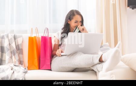 Une dame aux cheveux sombres vêtue d'un pantalon de survêtement confortable aux couleurs claires et un t-shirt souriant mystérieusement dans un ordinateur portable portant une carte de débit dans sa main, couvre-la Banque D'Images