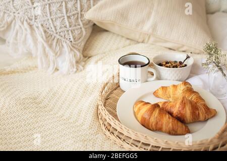 Gros plan sur plateau avec tasse à café, croissants, muesli et fleurs blanches sur couverture tricotée à la chambre douillette de style boho. Petit déjeuner, séjour lent, jom Banque D'Images