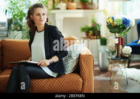femme pensive tendance d'âge moyen avec chemisier blanc et veste noire avec ordinateur portable fermé assis sur un canapé dans une maison moderne en journée ensoleillée. Banque D'Images
