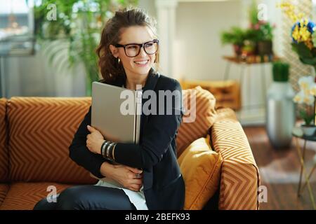 heureuse femme de 40 ans avec chemisier blanc et veste noire dans le salon moderne en journée ensoleillée assis sur canapé et un ordinateur portable près de la huggage. Banque D'Images