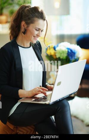 heureuse jeune femme dans un chemisier blanc et une veste noire à la maison moderne en journée ensoleillée avec ordinateur portable tout en étant assise sur un canapé. taper sur le clavier Banque D'Images