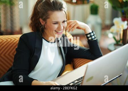 femme de ménage souriante et élégante avec chemisier blanc et veste noire dans le salon moderne en journée ensoleillée, message dactylographiant sur un ordinateur portable en étant assis Banque D'Images