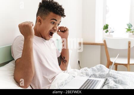Photo de jeunes hurlant surpris positif africain gars dans la chambre à coucher en utilisant ordinateur portable dans le lit faire le geste de winer. Banque D'Images