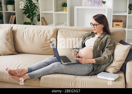 Jolie jeune femme enceinte dans la maison assise sur le canapé et lire l'article sur l'ordinateur portable tout en prenant des notes sur la maternité Banque D'Images